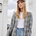 Erin wearing a plaid newsboy cap, plaid long jacket, plain white tshirt, and light wash blue jeans. Her hair is down and she is carrying a large black purse while standing in front of a large white building.
