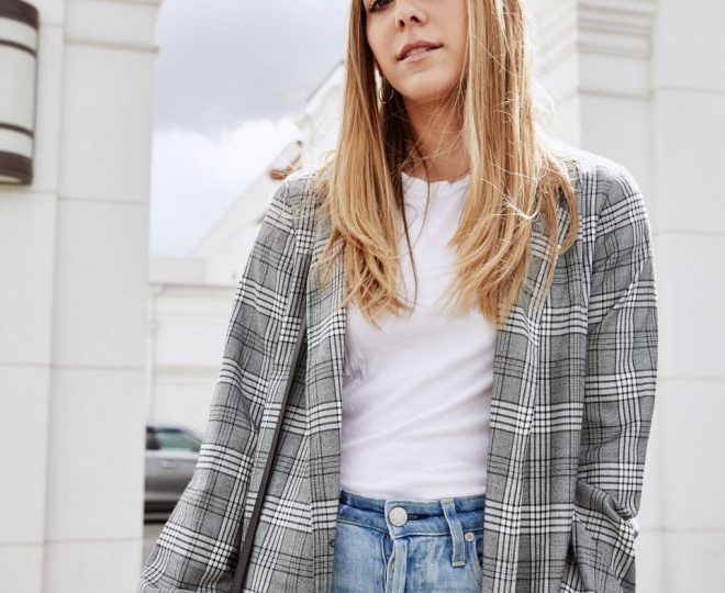 Erin wearing a plaid newsboy cap, plaid long jacket, plain white tshirt, and light wash blue jeans. Her hair is down and she is carrying a large black purse while standing in front of a large white building.