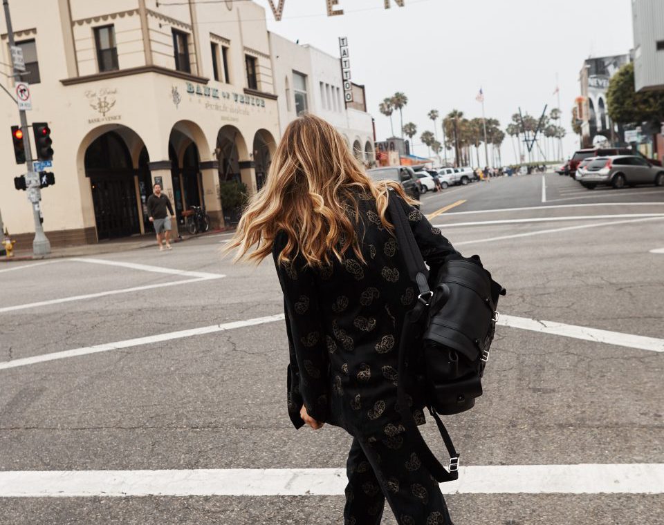 Erin crossing the street and wearing a patterned black suit with white sneakers and a black leather backpack