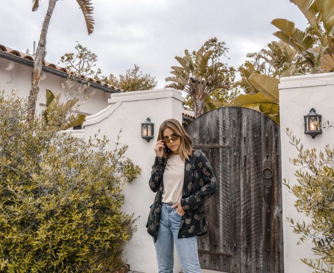 Erin in a patterned blazer and light blue jeans looking over the top of her sunglasses.