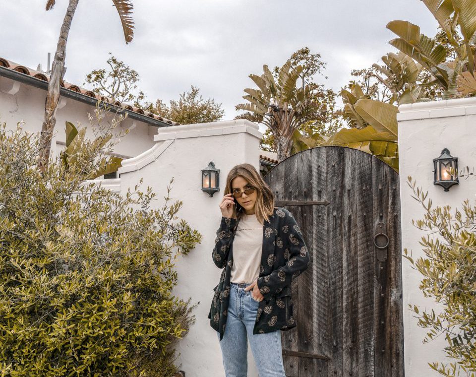 Erin in a patterned blazer and light blue jeans looking over the top of her sunglasses.