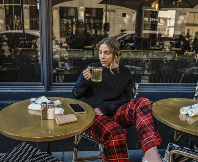Erin sitting at a cafe table drinking tea and wearing a black long sleeve shirt and red plaid pants with pink high heels.
