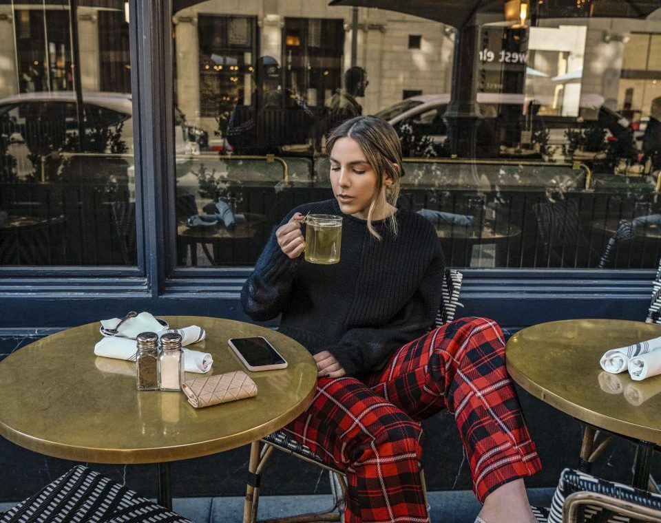 Erin sitting at a cafe table drinking tea and wearing a black long sleeve shirt and red plaid pants with pink high heels.