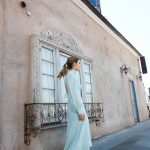 Erin wearing a blue flowing dress and black boots, walking in front of an ornate window and facing forward.