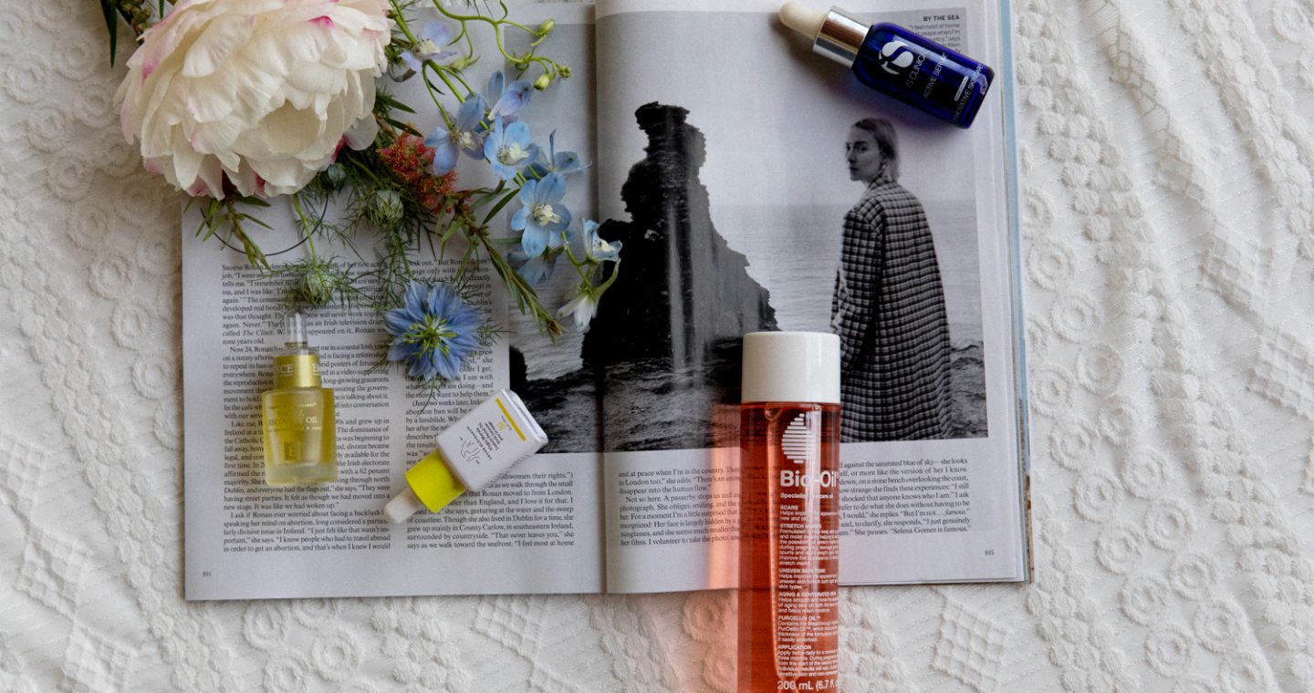 Bottles of face oil and a bouquet of flowers on top of an open magazine sitting on a knitted blanket