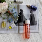 Bottles of face oil and a bouquet of flowers on top of an open magazine sitting on a knitted blanket