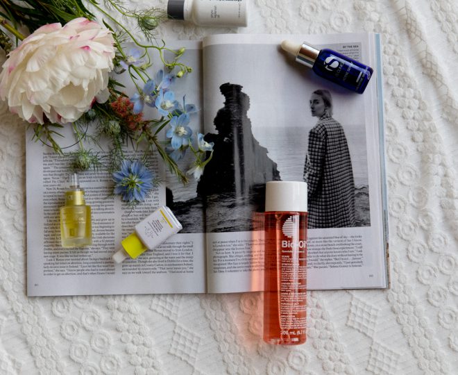 Bottles of face oil and a bouquet of flowers on top of an open magazine sitting on a knitted blanket