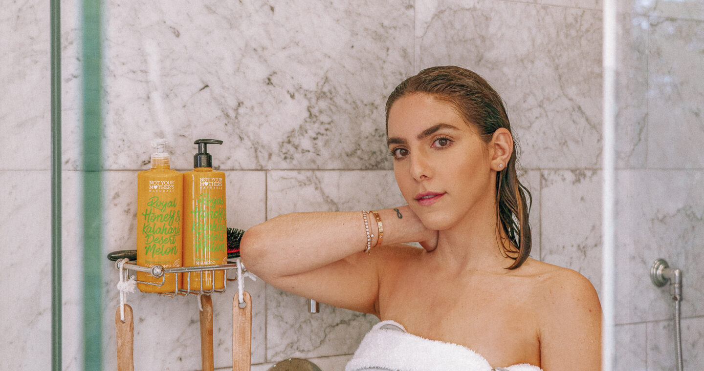 Erin standing in the shower with a white towel wrapped around her. Not Your Mother's shampoo is in the background.