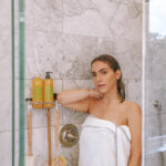 Erin standing in the shower with a white towel wrapped around her. Not Your Mother's shampoo is in the background.