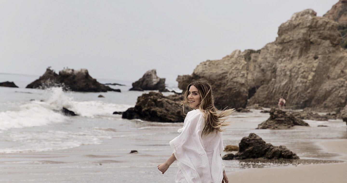 Erin walking along the beach in a white cover up, looking back over her shoulder.