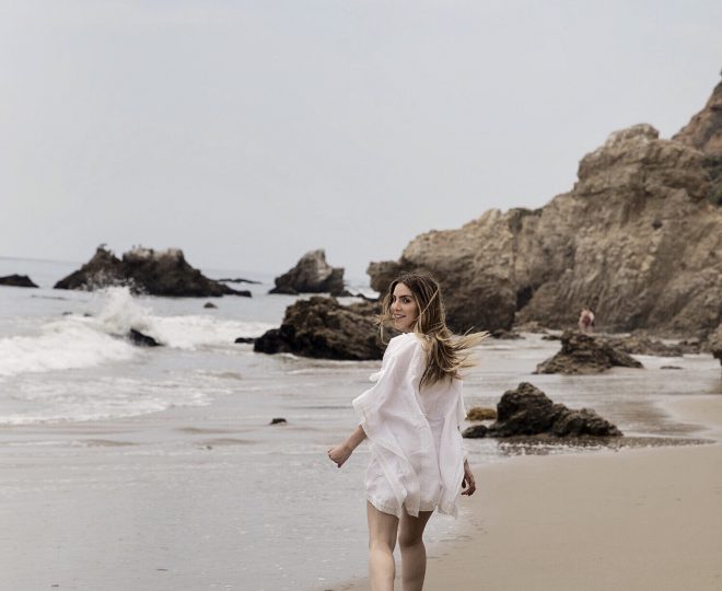 Erin walking along the beach in a white cover up, looking back over her shoulder.